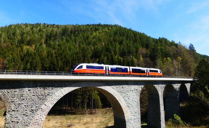 A train going over a stone bridge in Austria, there is a hill covered in green pines on the background. An example of the journeys on this London to Vienna Train Tour.