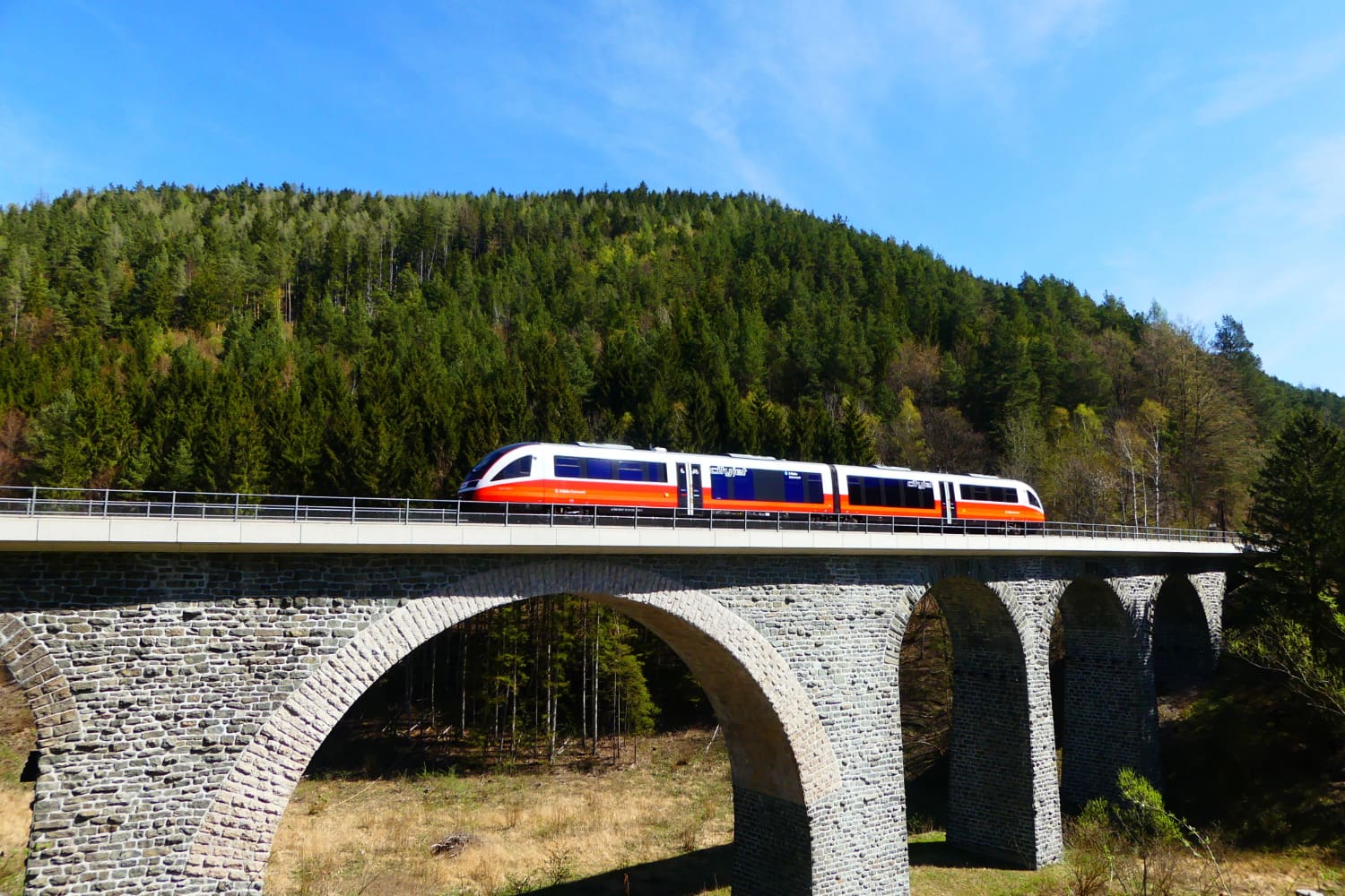 A train going over a stone bridge in Austria, there is a hill covered in green pines on the background. An example of the journeys on this London to Vienna Train Tour.